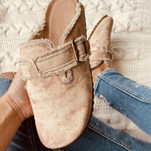 Beige slip-on shoe with a buckle held by a person wearing blue jeans on a textured white background.