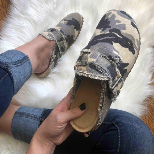 Camouflage-patterned slip-on shoes being held by a person wearing jeans on a white fur surface.