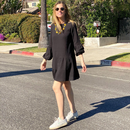 A woman is standing on the sidewalk wearing a black mini dress with puffed sleeves and a tiered ruffled mini hem, paired with sunglasses and white shoes.