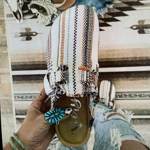 A person holds a white wedge slide sandal with vertical black, brown, and orange stripes and frayed fringe detailing. The sandal has a tan footbed with a silver script logo.