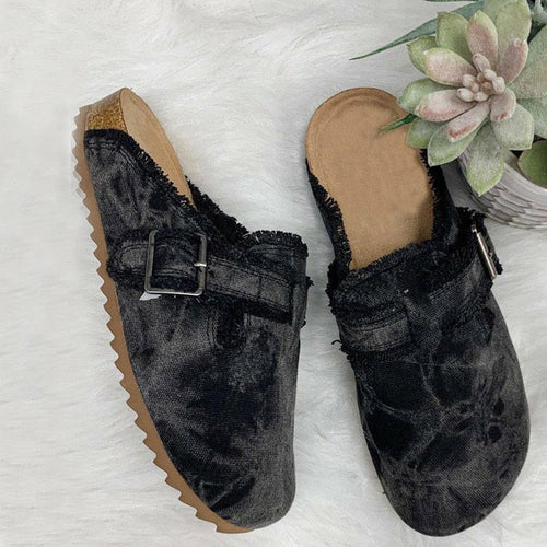 A pair of women's mule clogs with a dark gray and black marbled or tie-dyed textured upper, a frayed edge, a wide strap with a silver buckle, and a brown cork-look sole, displayed on a white faux fur background with a small succulent plant.