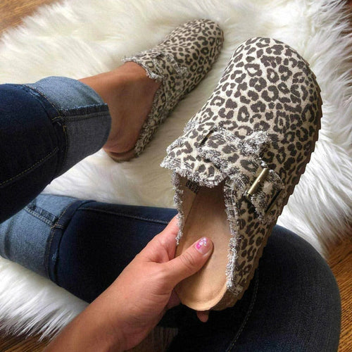 leopard-print slip-on clogs with frayed trim and a decorative strap, sitting on a white faux-fur rug. One shoe is on the foot, and the other is being held by a hand to show the interior.