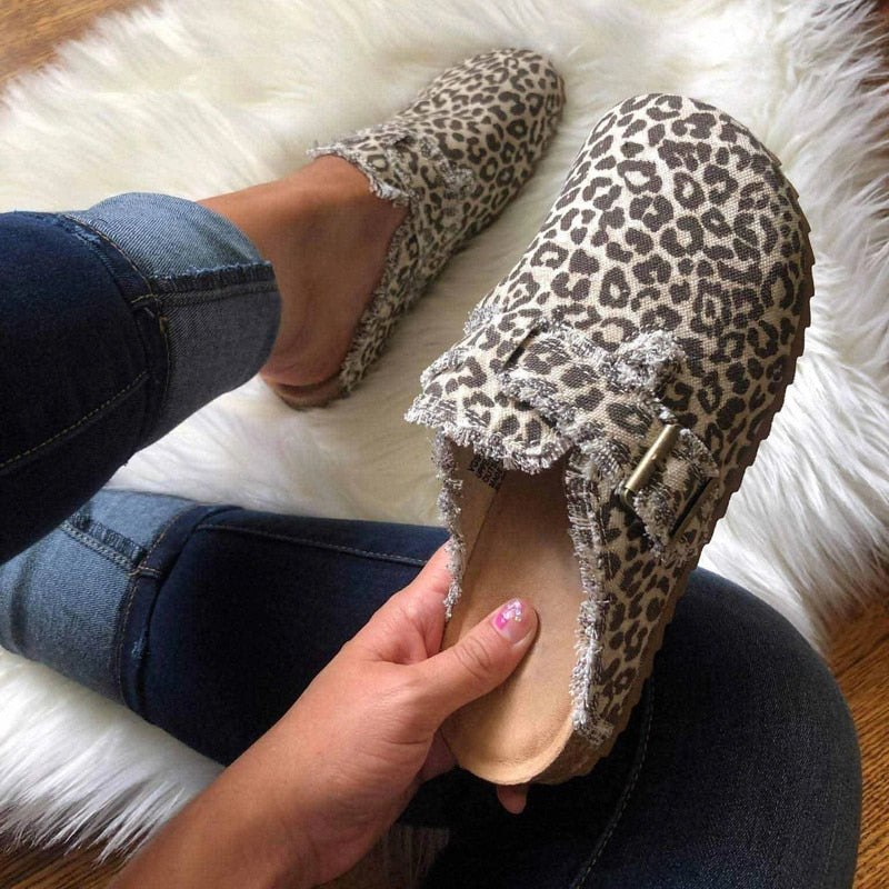 A person wearing leopard-print slip-on clogs with frayed trim and a decorative strap, sitting on a white faux-fur rug. One shoe is on the foot, and the other is being held by a hand to show the interior.